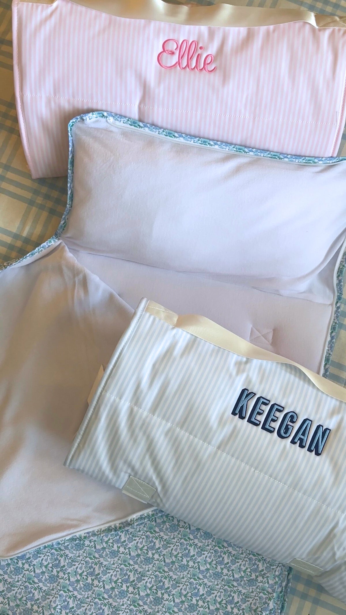 Three personalized Nap Mats in pink, blue, and floral prints, embroidered with the names "Ellie" and "Keegan." The mats are soft, padded, and feature built-in pillows, perfect for preschool or daycare naptime. One mat is fully unrolled to showcase its cozy interior and quilted design.