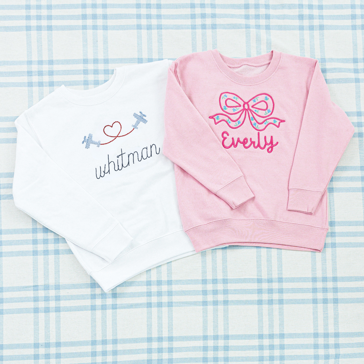 Personalized Crewneck Sweatshirts with Full Chest Motif from Rainey Laine, featuring a white sweatshirt with embroidered airplanes forming a heart and the name ‘Whitman,’ and a pink sweatshirt with an oversized bow motif and the name ‘Everly,’ displayed on a soft blue plaid background.