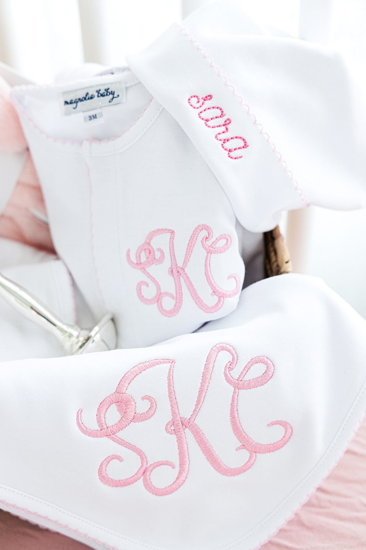 The image shows a white baby set with pink embroidery, featuring a monogrammed "SK" on a blanket and the name "Sara" on a beanie. 