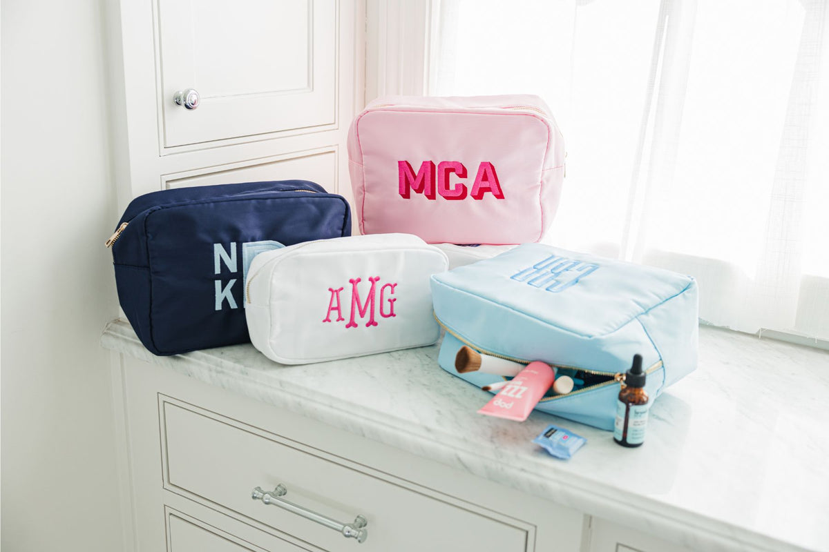 A collection of personalized pouches, including a navy pouch, a white pouch, a pink pouch, and a light blue pouch.