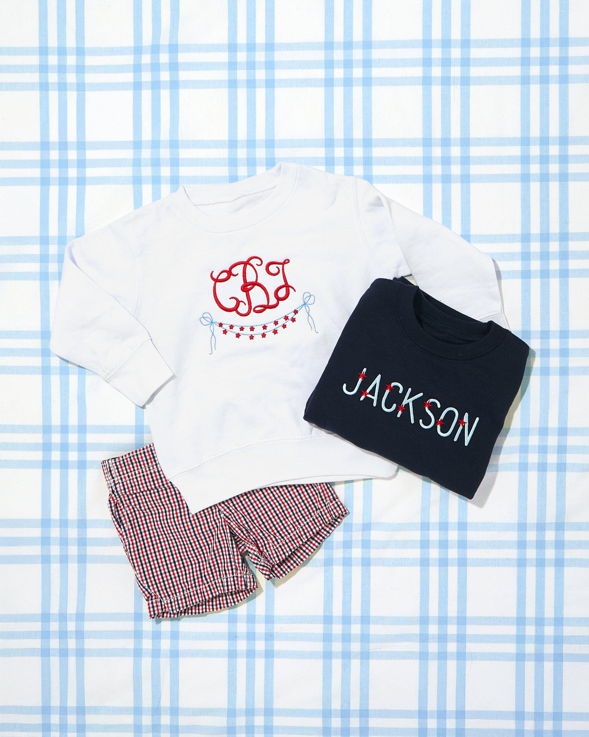 Flat lay featuring two personalized Crewneck Sweatshirts with Full Chest Name embroidery—one in white with a monogram and patriotic bunting motif, and the other in navy with “JACKSON” boldly stitched in red, white, and blue. Paired with coordinating red, white, and blue gingham shorts, creating a festive and cozy look perfect for chilly summer evenings or patriotic celebrations.