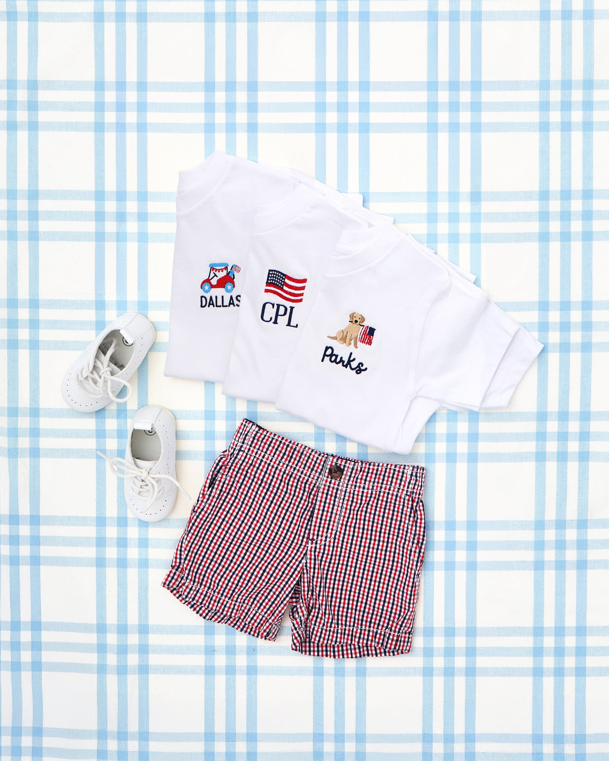 Flatlay of three Solid Short Sleeve T-Shirts with Motif, each featuring patriotic-themed embroidery and personalized names: a red scooter with “DALLAS,” an American flag with initials “CPL,” and a golden retriever pup holding a flag with “Parks.” Paired with red, white, and blue gingham shorts and white lace-up shoes, this festive ensemble is perfect for 4th of July celebrations or summer outings. Set against a pastel plaid background for a charming Americana vibe.