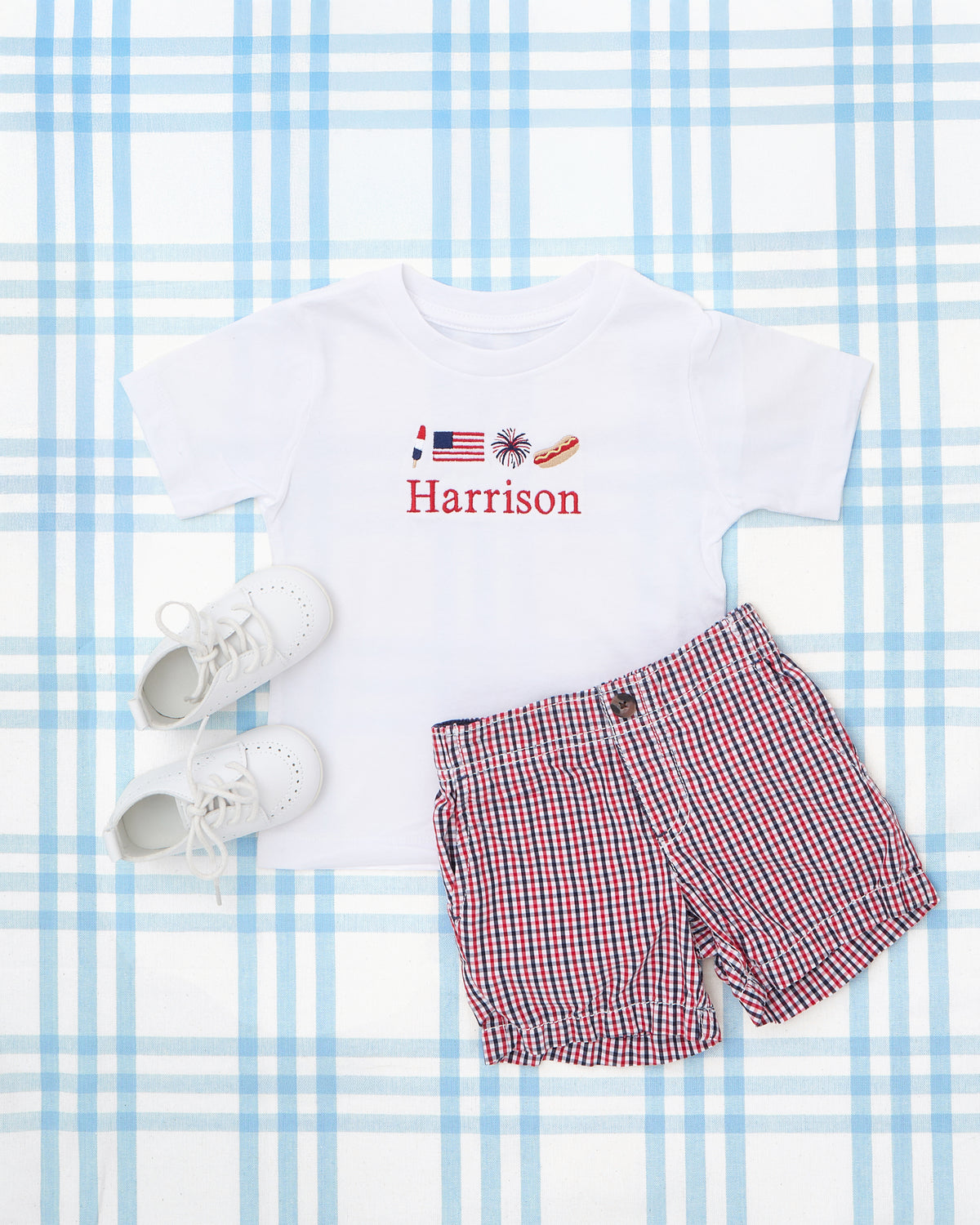 Flat lay of a personalized baby boy’s outfit featuring a white Short Sleeve T-Shirt with Full Chest Patriotic Motif, embroidered with a popsicle, American flag, fireworks, hotdog, and the name “Harrison” in bold red text. Paired with red, white, and blue gingham shorts and classic white baby shoes—perfect for festive summer celebrations like the 4th of July.