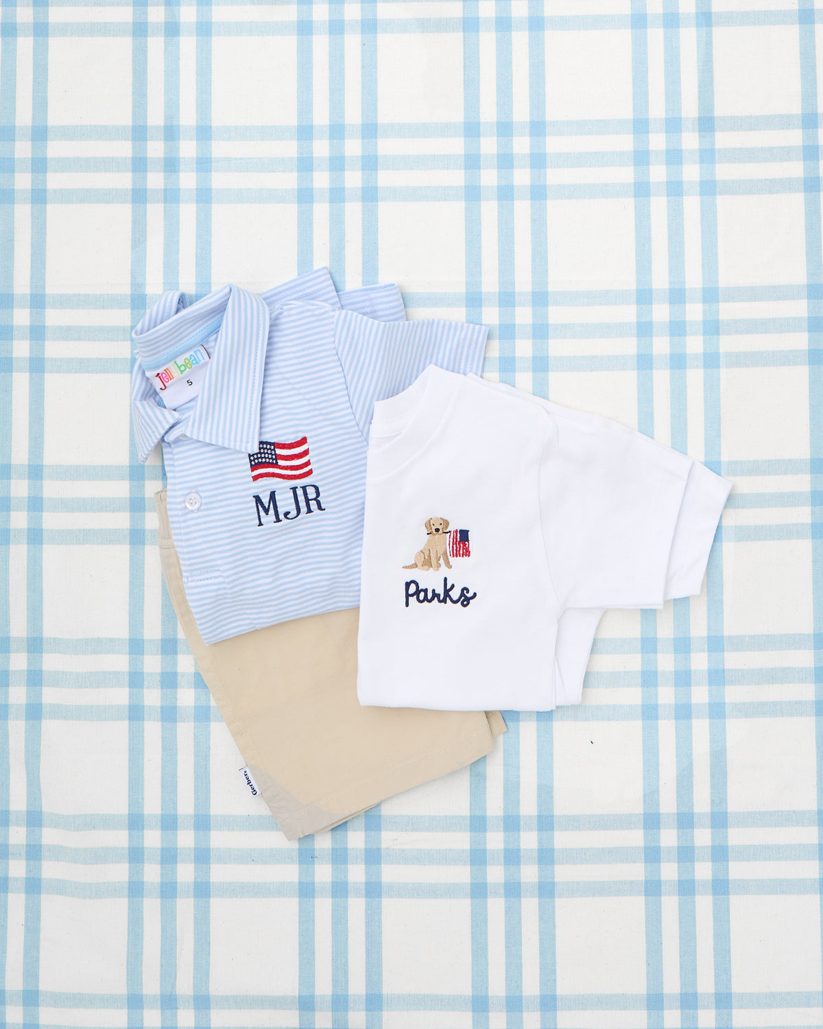 Personalized Solid Short Sleeve Polo Shirts for boys, featuring embroidered patriotic motifs—one light blue striped polo with an American flag and initials "MJR," and one classic white tee with a golden retriever holding an American flag and the name "Parks." These charming tops pair perfectly with khaki bottoms for a polished, all-American look.