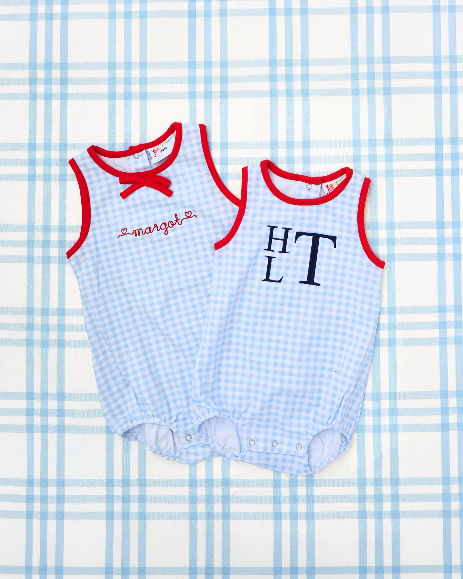 Two Blue & Red Gingham Bubbles displayed on wooden hangers. The sleeveless baby bubbles feature a light blue gingham print with bold red piping around the armholes and neckline. One style is accented with a red embroidered name "Barrett," while the other features a red bow detail and black monogram "EMB," making them perfect for coordinating sibling outfits with a patriotic or preppy twist.