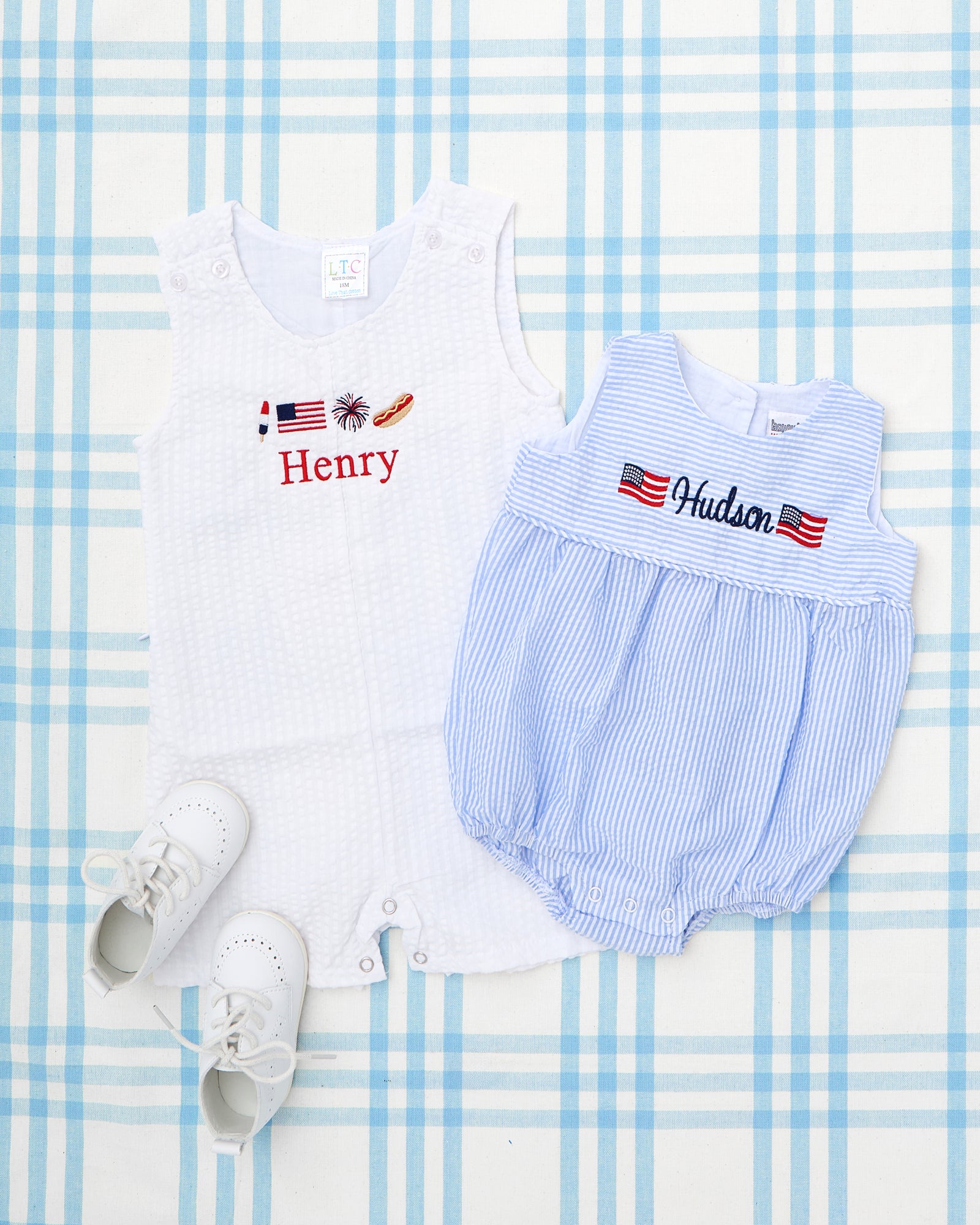 Two personalized baby seersucker bubbles are shown side by side—one in blue and white stripes with the name "Hayden" embroidered between two American flags, and the other in pink and white stripes with the name "Sophia" embroidered between two pink tulip motifs. These Seersucker Bubbles with Motif feature a classic sleeveless silhouette, soft gathers at the waist, and custom embroidery for a timeless, preppy look.