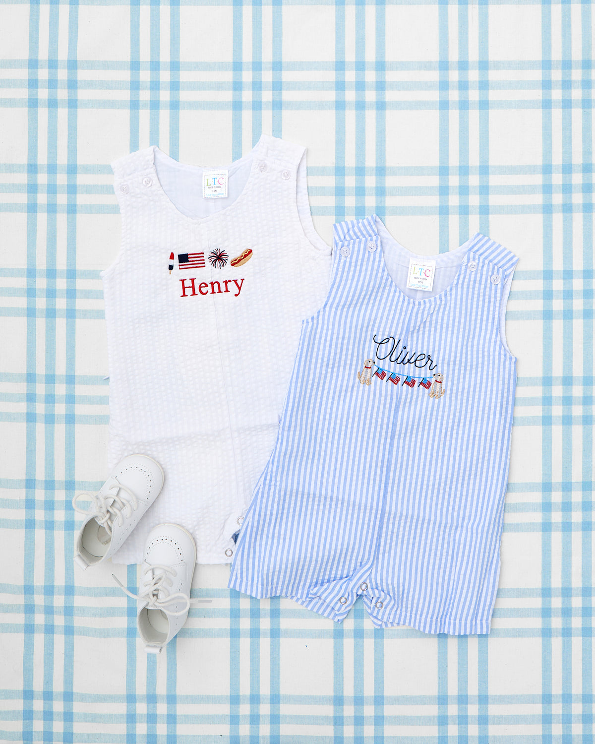 Two Seersucker Jon Jons with Patriotic Motifs are laid out on a blue plaid backdrop, paired with classic white baby shoes. The white Jon Jon features embroidered patriotic icons—including a popsicle, American flag, firework, and hotdog—above the name “Henry” in bold red. The blue-striped jon jon showcases a nautical design with life preservers and bunting flags, embroidered above the name “Olivier” in navy script—a perfect set for festive summer celebrations, such as the Fourth of July.