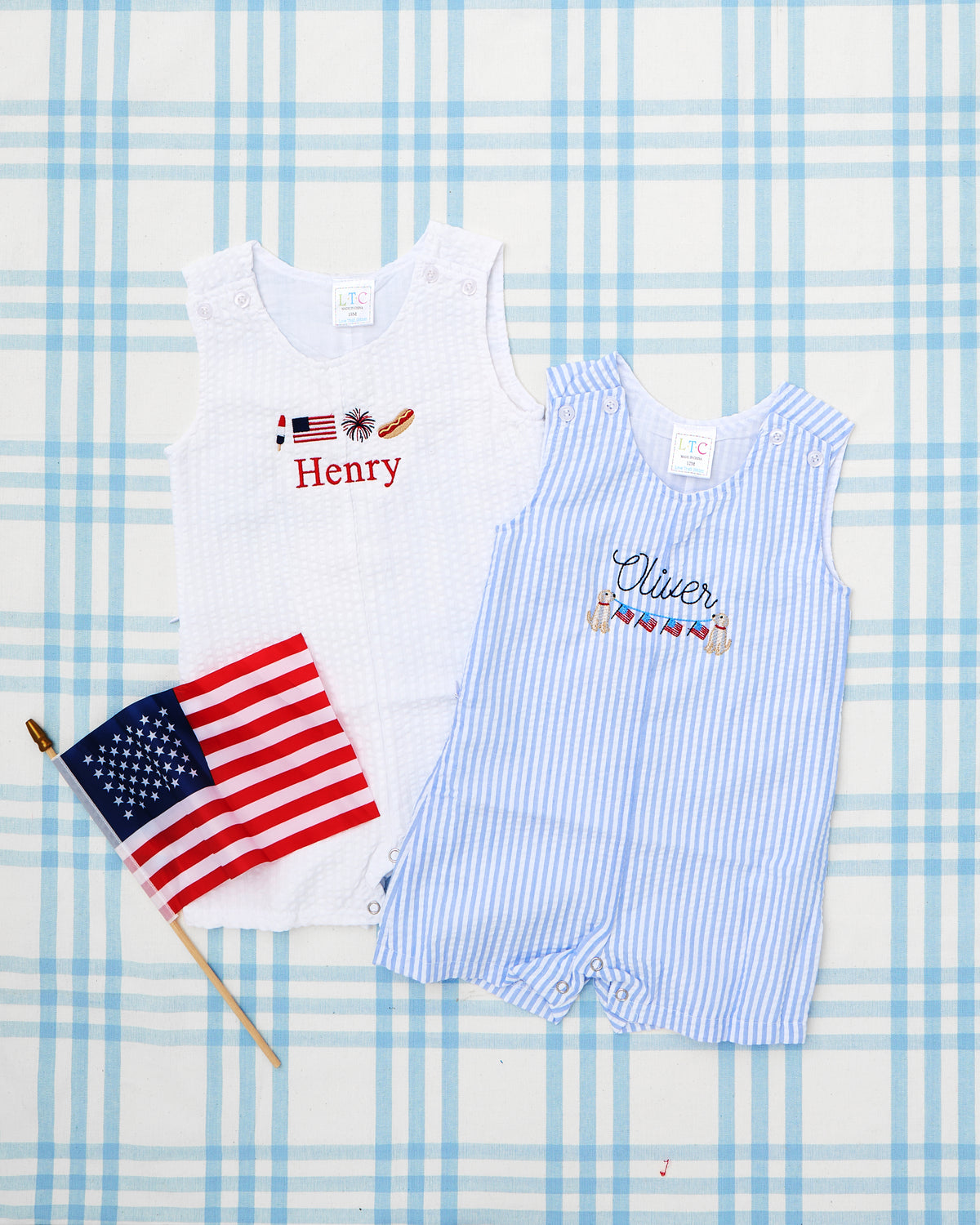 Two Seersucker Jon Jons with Patriotic Motifs displayed on a soft blue plaid background. The white jon jon features festive embroidery with a flag, firework, and hotdog above the name “Henry” in bold red thread. The blue-and-white striped jon jon showcases a nautical bunting motif with American flags and life preservers above the name “Olivier” in navy script. Both outfits embody classic American summer charm—perfect for 4th of July celebrations and family festivities.
