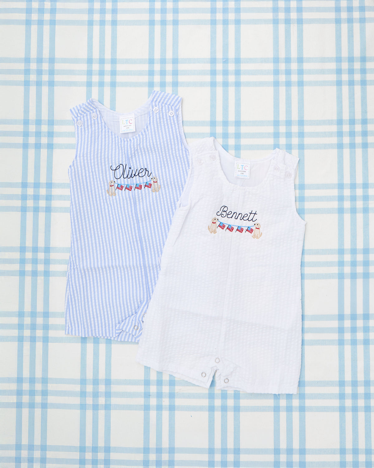 Flatlay of two personalized Seersucker Jon Jons with Patriotic Motif featuring sleeveless designs in classic blue and white seersucker fabric. Each romper is custom-embroidered with a name—"Oliver" on the blue stripe and "Bennett" on the white—with a festive banner of American flags held by playful golden retriever puppies. Perfect for celebrating summer holidays like the Fourth of July in timeless Southern style. Displayed on a light blue plaid background.