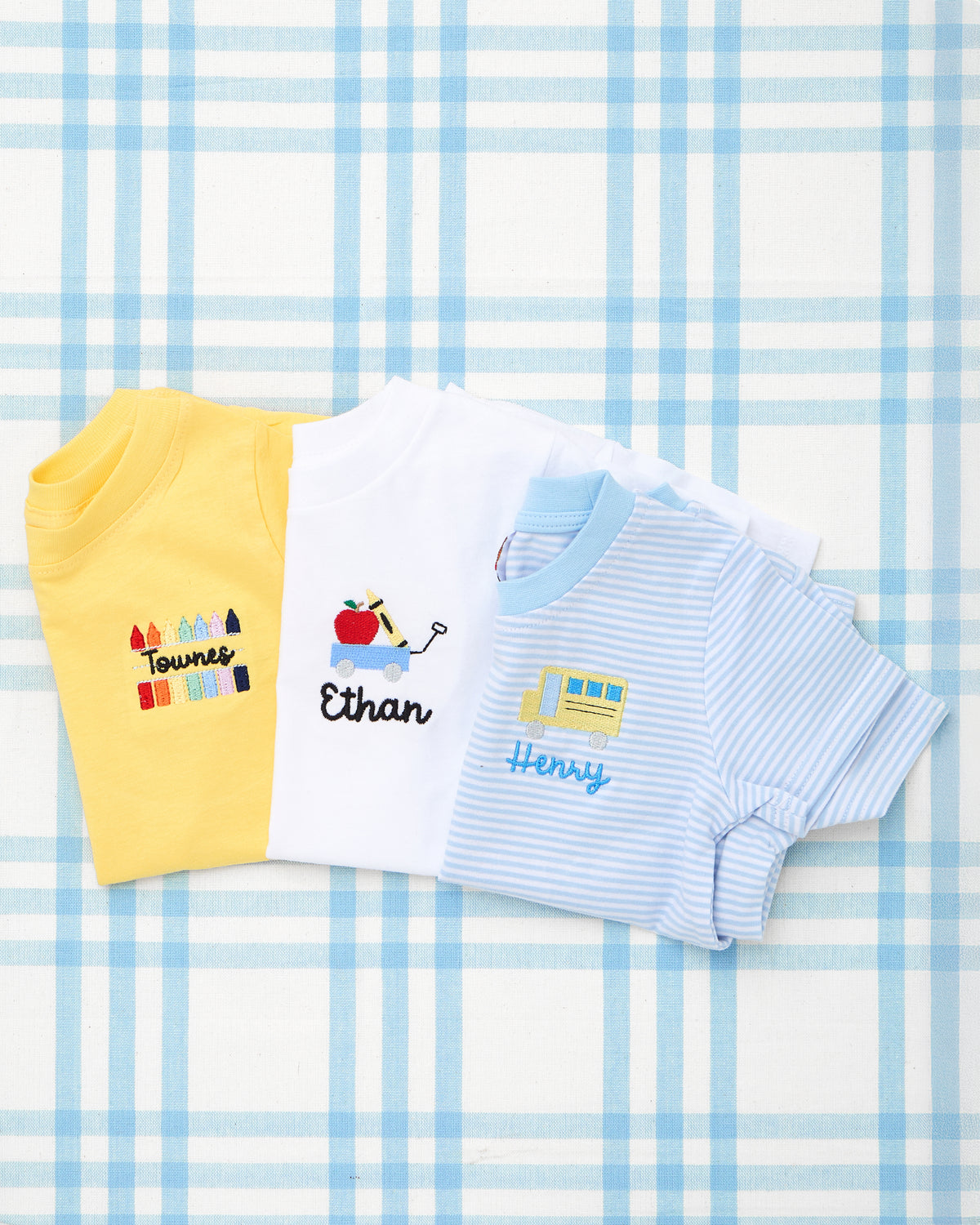 Set of three personalized Solid Short Sleeve T-Shirts with Motif, featuring playful school-themed embroidery: a yellow tee with crayon motif and "Townes," a white tee with a wagon of school supplies and "Ethan," and a light blue striped tee with a yellow school bus and "Henry"—perfect for back-to-school outfits with a personalized touch.