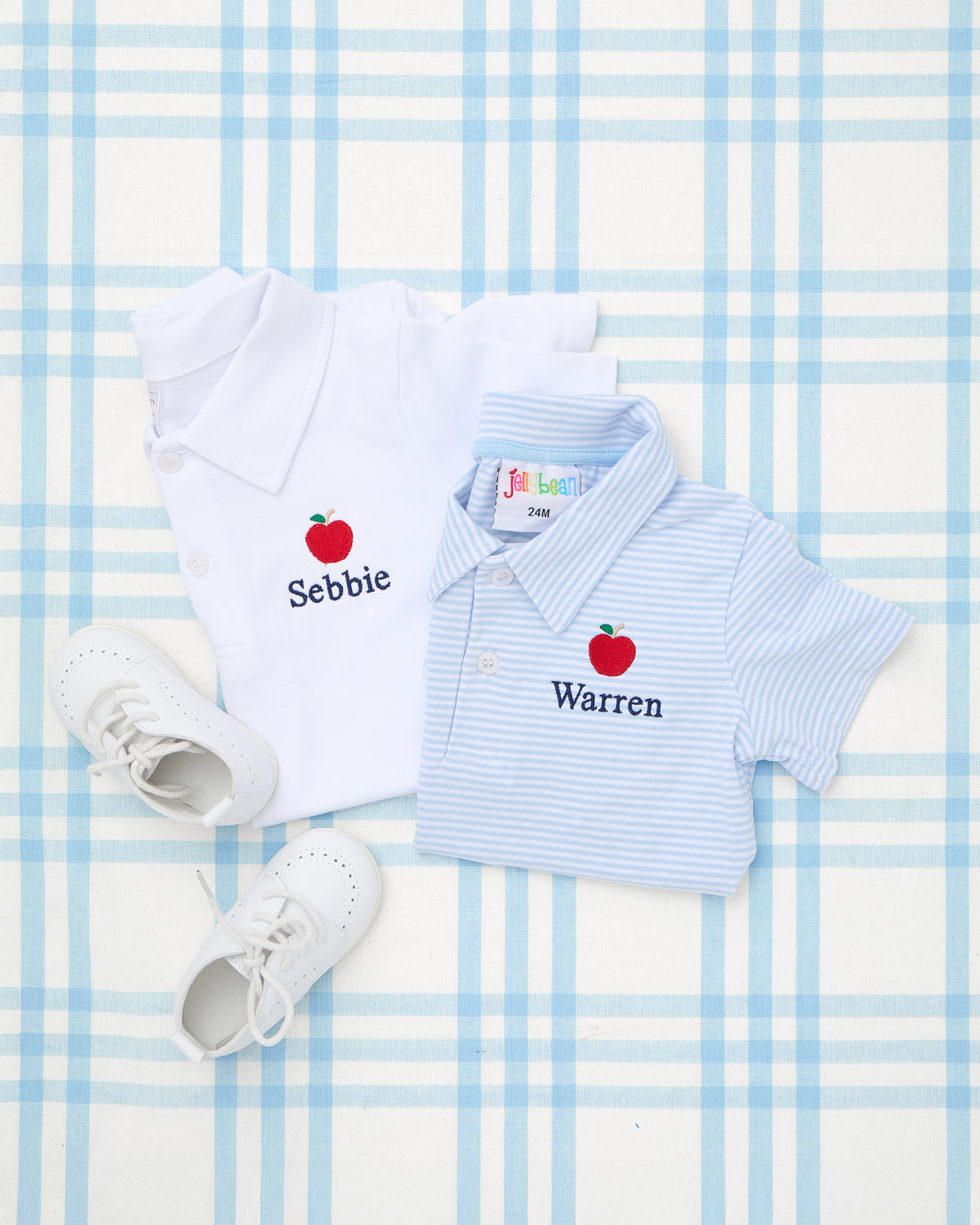 Two personalized Solid Short Sleeve Polos with Motif, featuring classic collar styles and embroidered red apple designs—one in solid white with the name "Sebbie," and the other in light blue stripes with the name "Warren." A charming back-to-school outfit perfect for celebrating the first day of school in style.