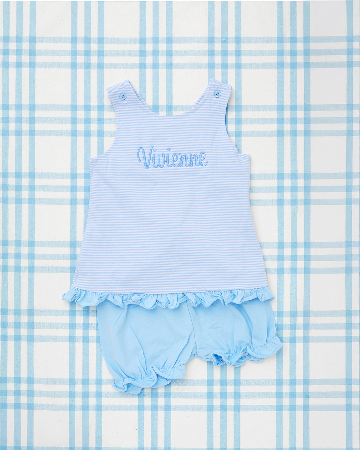 Blue and white Striped Criss Cross Bloomer Set for toddlers, featuring a sleeveless top with subtle horizontal stripes, ruffle hem detailing, and the name "Vivienne" embroidered in coordinating blue thread. The matching bloomers have elastic leg openings with ruffle accents, creating a sweet and classic silhouette perfect for warm-weather outings or everyday play. Set is displayed on a blue plaid background.