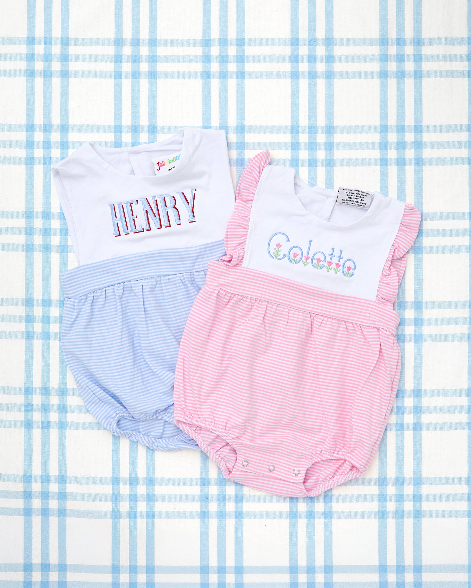 Two sleeveless striped bubbles for babies are displayed on wooden hangers—one in light blue with the name "Calvin" embroidered in playful green starfish lettering, and the other in light pink with ruffle sleeves and monogram "EMG" in a classic font. These Sleeveless Striped Bubbles feature soft horizontal pinstripes, snap closures at the bottom, and personalized embroidery, perfect for summer days or special occasions.