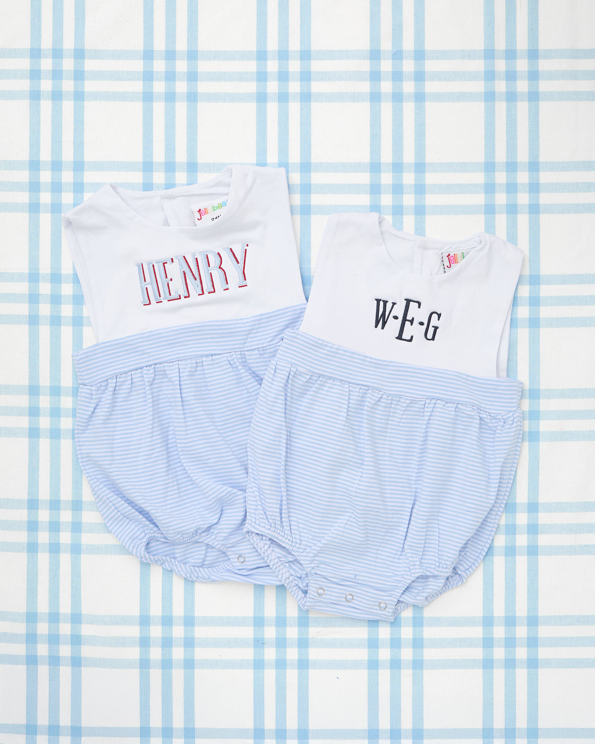 Two Sleeveless Striped Bubbles for toddlers featuring white bodices with custom embroidered names and light blue pinstriped bubble bottoms. One bubble displays the name “HENRY” in bold red outlined text, while the other features the initials “W•E•G” in classic black serif font. Both garments have gathered elastic leg openings and snap closures, designed for a comfortable and polished look ideal for warm weather. Set is laid on a blue plaid backdrop.