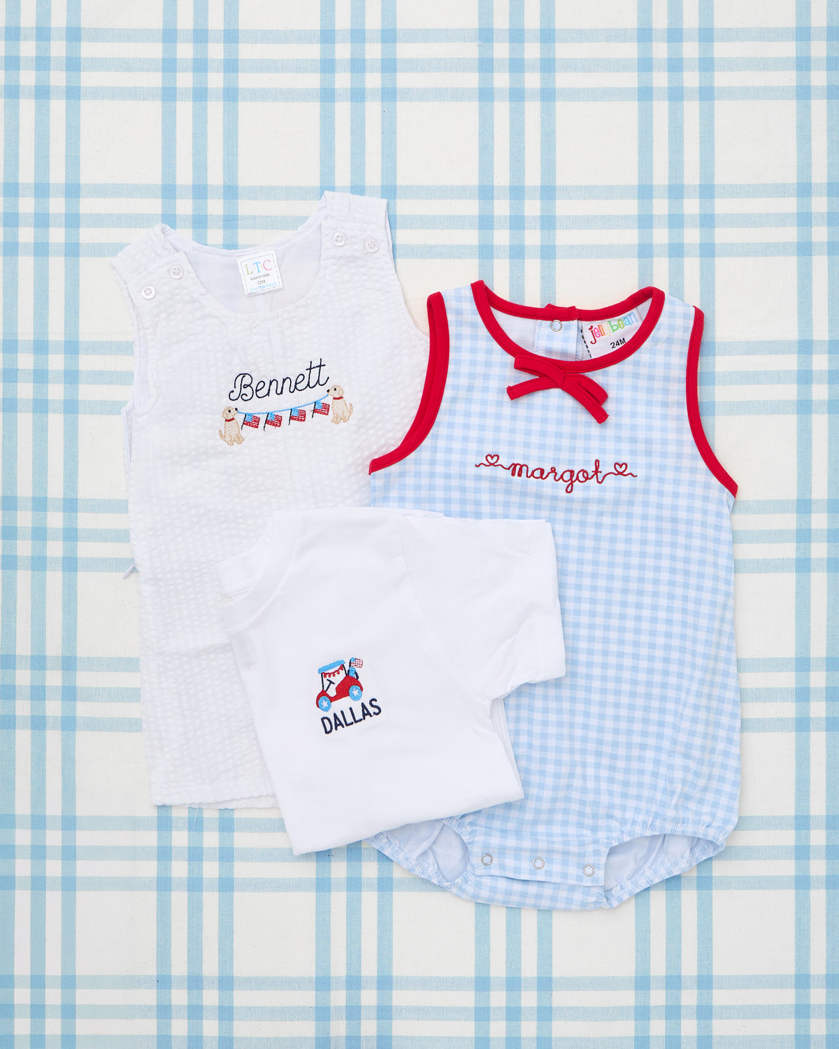 Flat lay featuring a patriotic-themed baby outfit set on a light blue plaid background. The right side highlights a Blue & Red Gingham Bubble with red piping, a decorative front bow, and the name “Margot” embroidered in red script with heart details. The ensemble also includes a white Seersucker Jon Jon with the name “Bennett” and dog holding flag garlands, and a white t-shirt folded to display a red scooter motif with the name “DALLAS” below it—perfect for festive summer celebrations like the Fourth of Jul