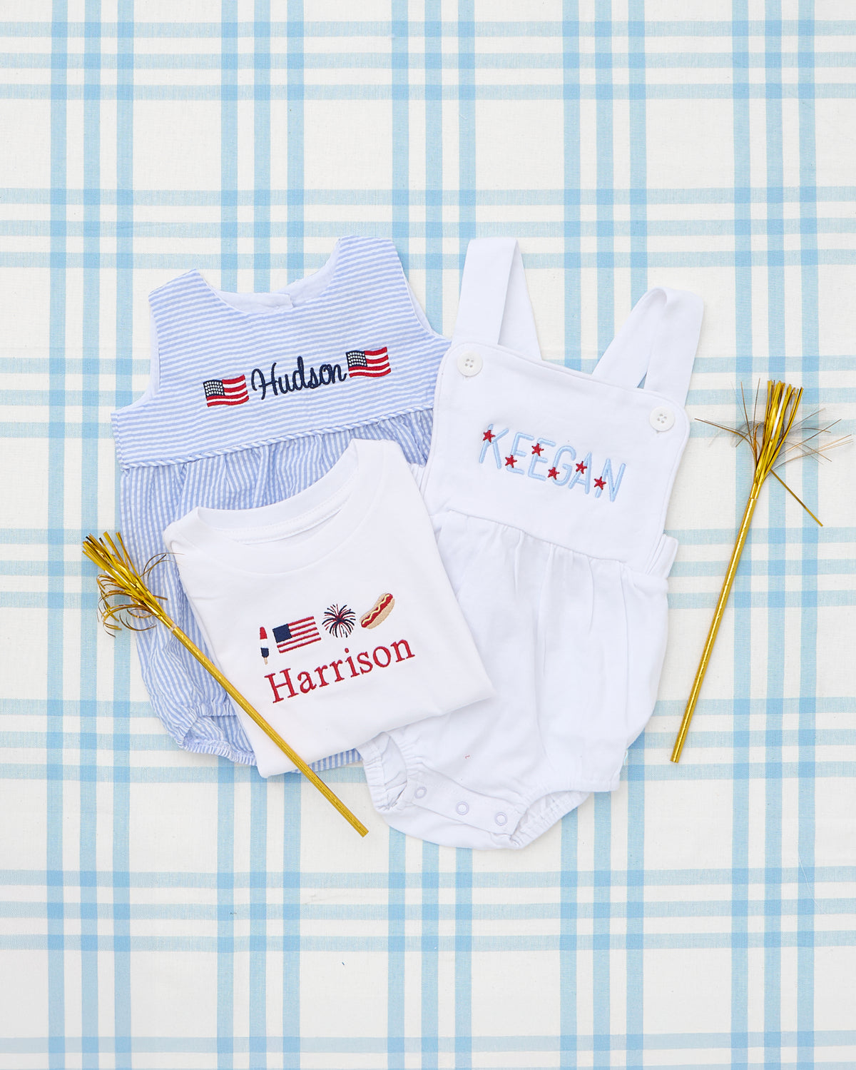 Flat lay featuring a trio of patriotic children's outfits on a blue plaid background. Highlighted is a white Cotton Sunsuit with “KEEGAN” embroidered in red, white, and blue stars. Alongside it is a striped Seersucker Bubble with flag motifs and “Hudson” stitched across the chest, and a white Short Sleeve T-Shirt with a full chest patriotic motif including a flag, firework, and hot dog, personalized with the name “Harrison.” Perfect for festive Fourth of July celebrations.