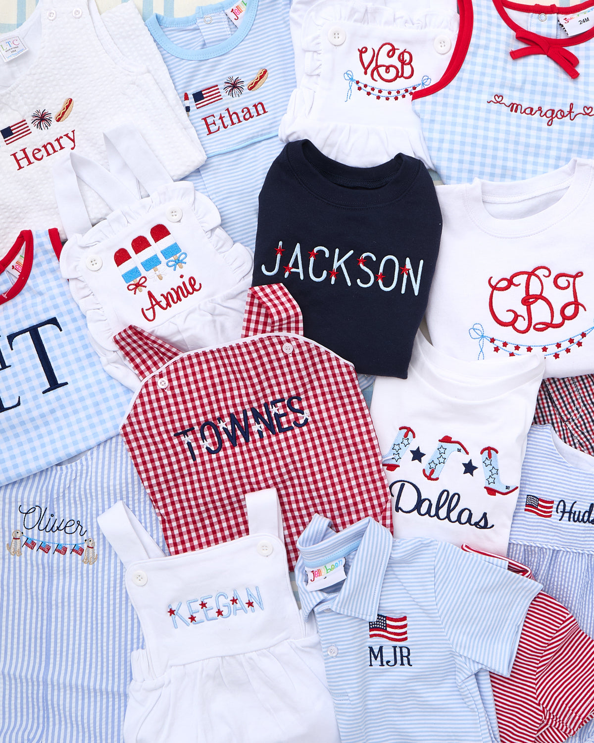 Flat lay of personalized children’s outfits featuring a variety of embroidered patriotic designs. Centered is a navy Crewneck Sweatshirt with Full Chest Patriotic Motif, customized with the name “JACKSON” in red, white, and blue lettering. Surrounding garments include jon jons, polos, sunsuits, and t-shirts with American flags, popsicles, and festive summer accents—perfect for celebrating Independence Day in style.