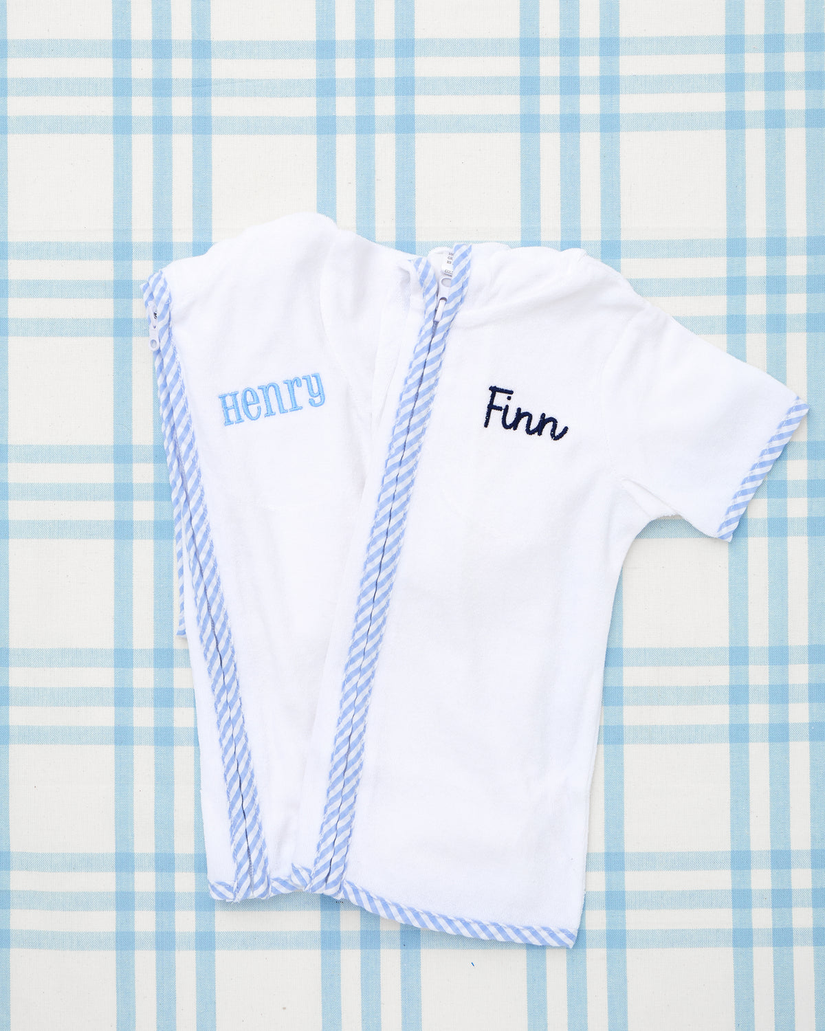 Two personalized kid’s Terry Towel Robes laid flat on a blue plaid background, each featuring soft white terry cloth material with light blue striped trim. One robe is embroidered with the name "Henry" in block letters, and the other with "Finn" in a script font—perfect for post-swim comfort or bath time.