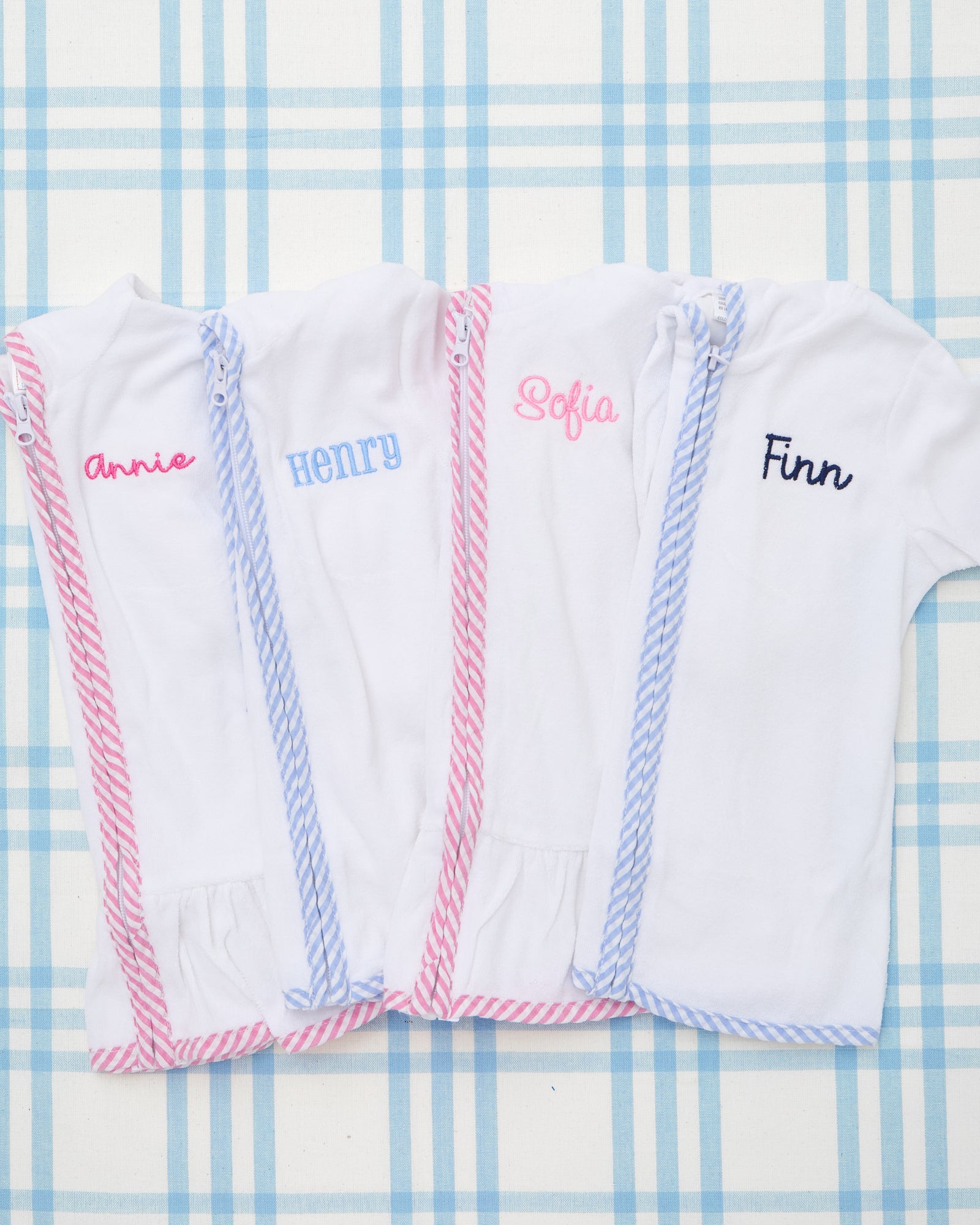Two Kid's Terry Towel Robes with hoods are displayed side by side—one with blue striped trim and the name "Beau" embroidered in a bold navy font, and the other with pink striped trim, ruffled hem, and the monogram "MJQ" stitched in soft pink. These lightweight robes are made from plush terry cloth, featuring short sleeves, zip-up fronts, and personalized embroidery, perfect for beach days, poolside drying, or cozy bath time.