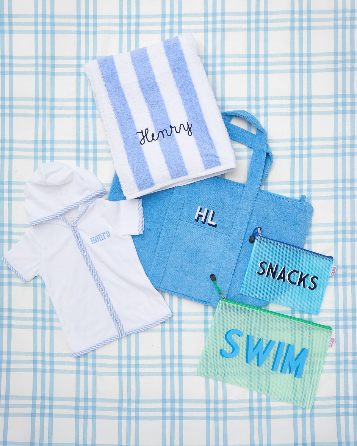 Flat lay featuring a personalized kid’s Terry Towel Robe with light blue striped trim and the name “Henry” embroidered on the chest, ideal for poolside comfort. The set also includes a matching blue and white striped beach towel, a monogrammed terry cloth tote bag with “HL,” and two mesh pouches labeled “SNACKS” and “SWIM” for organized summer essentials. Perfect for a day at the beach or pool.