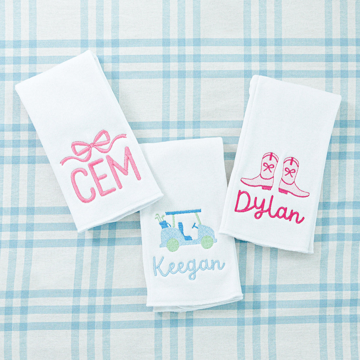 Three personalized towels with embroidered names: 'CEM' with a pink bow, 'Keegan' with a golf cart, and 'Dylan' with cowboy boots.