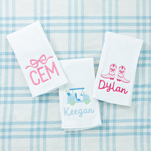 Three personalized towels with embroidered names: 'CEM' with a pink bow, 'Keegan' with a golf cart, and 'Dylan' with cowboy boots.