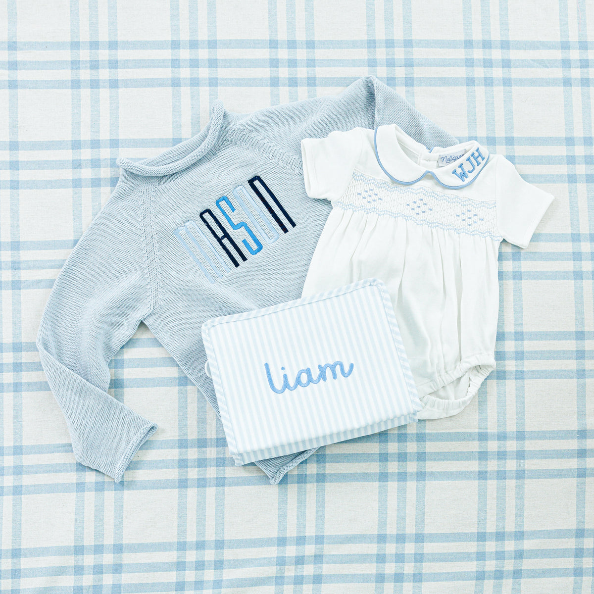 A charming baby gift set featuring a personalized blue sweater, a white onesie and a pouch.