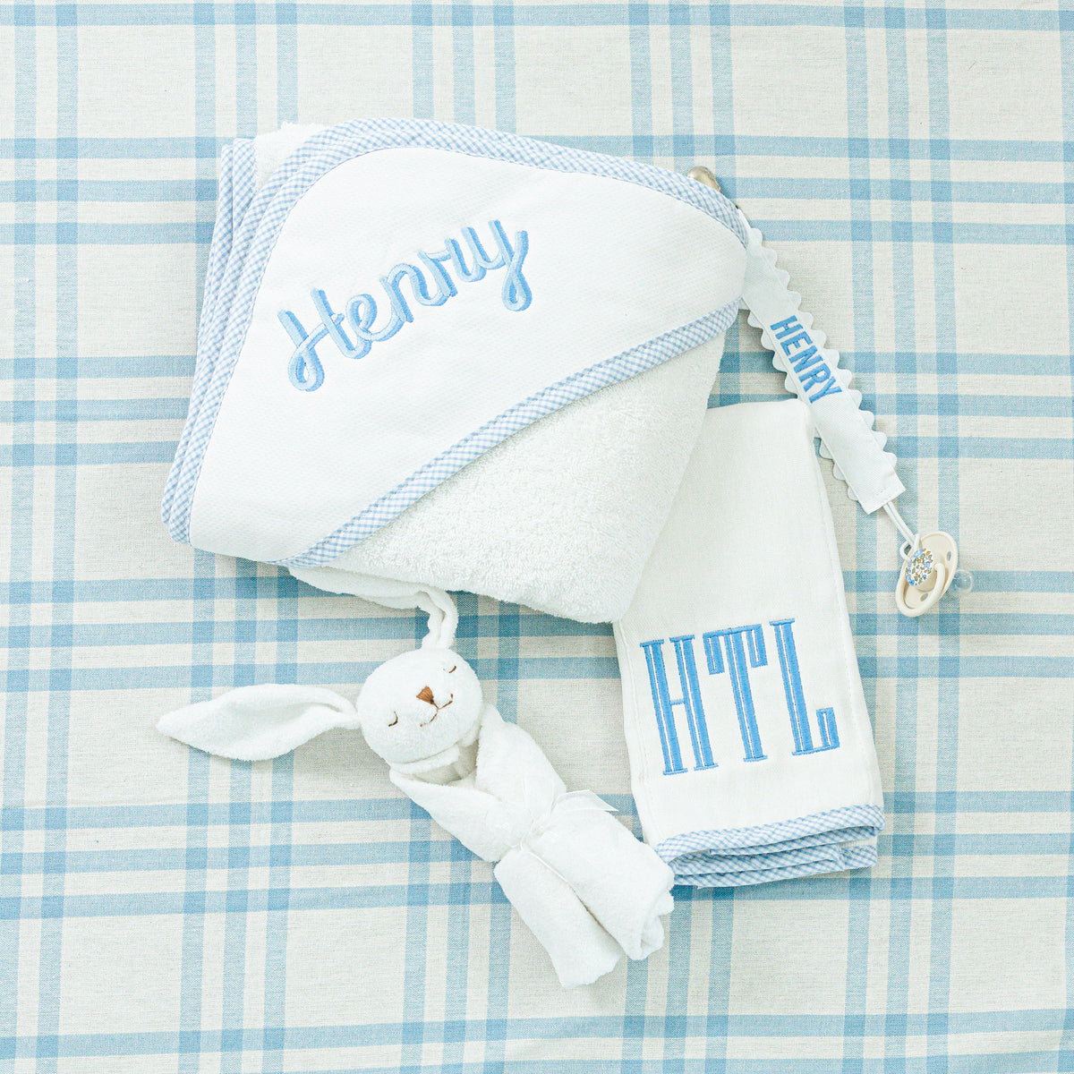 A personalized baby gift set displayed on a blue plaid background, featuring a white hooded towel with the name 'Henry' embroidered in blue, a matching white burp cloth with the initials 'HTL,' and a pacifier clip also embroidered with 'Henry,' accompanied by a soft bunny plush.