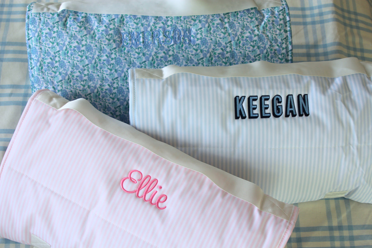 Three personalized Nap Mats rolled up and embroidered with names—Emerson, Keegan, and Ellie—featuring soft floral, blue striped, and pink striped designs. These cozy, all-in-one mats include a built-in pillow and blanket, ideal for daycare, preschool, or naptime on the go.