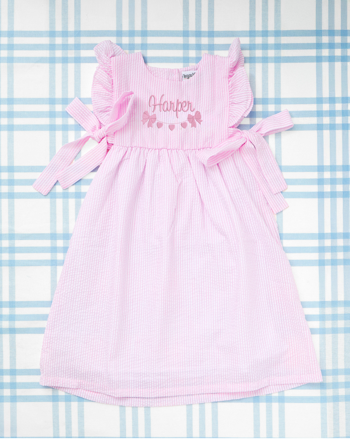 Seersucker Bow Dress with Motif from Rainey Laine, featuring pink striped fabric, ruffled sleeves, and side tie bows, with a personalized embroidered name ‘Harper’ above a pink bow and heart garland motif, displayed on a soft blue plaid background.