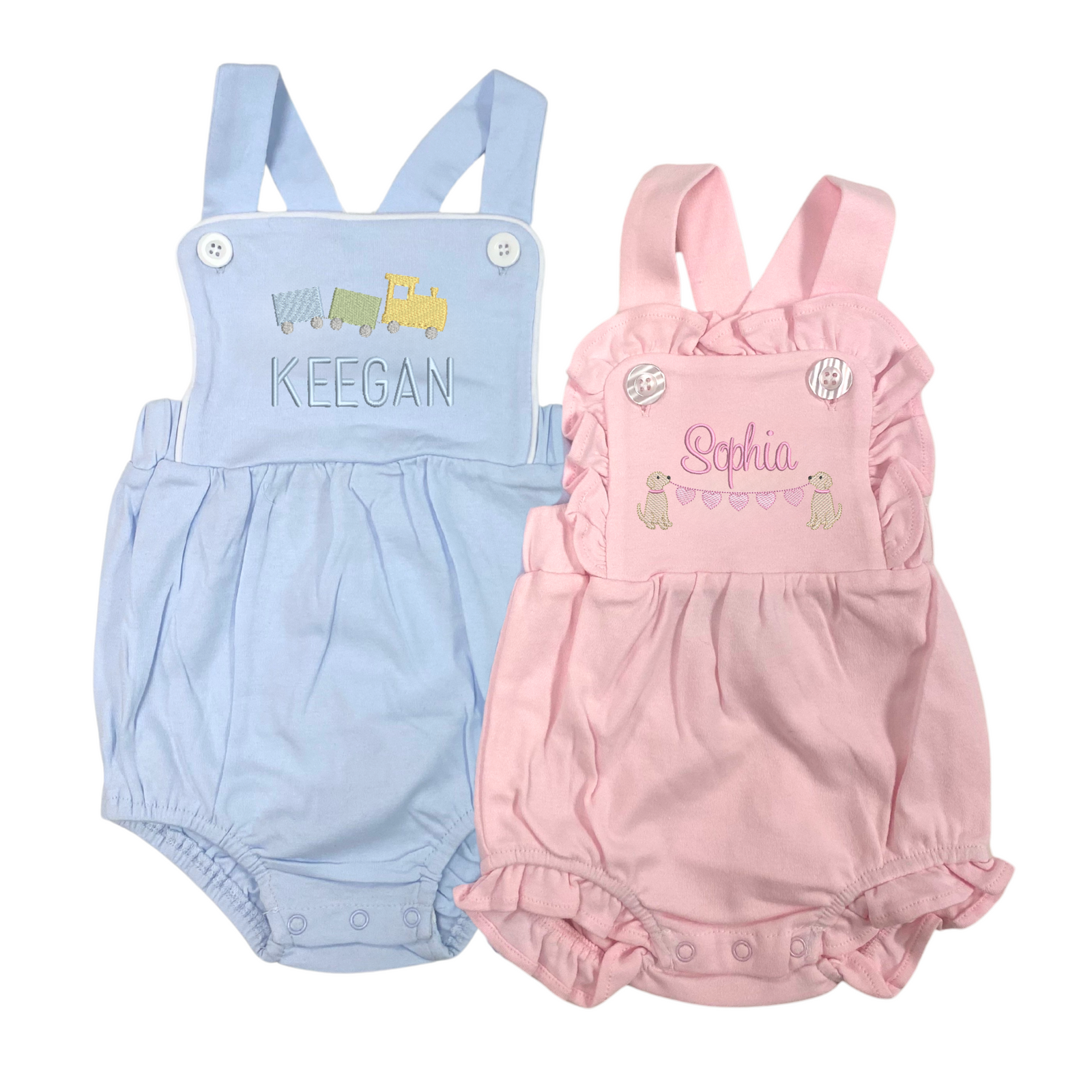 Image of two Cotton Sunsuits with Motif for toddlers—one in light blue and one in soft pink. The blue sunsuit features embroidered train cars above the name "KEEGAN," while the pink version showcases adorable puppy motifs and a string of hearts above the name "Sophia." Both sunsuits have shoulder button straps and elastic leg openings for a comfy, playful fit.