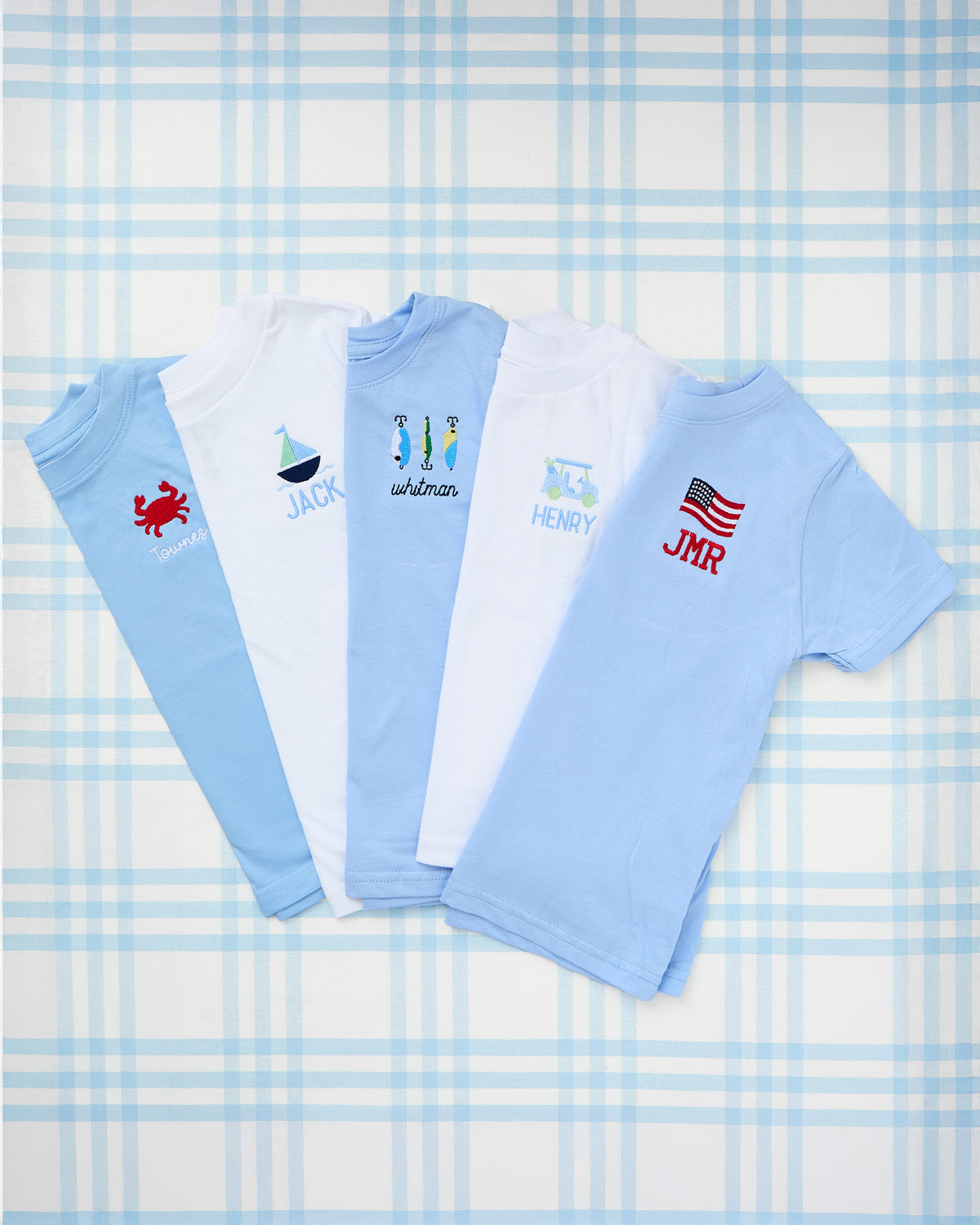 Flat lay of personalized kids' solid short sleeve t-shirts with embroidered motifs including a crab, sailboat, fishing lures, tractor, and American flag—perfect for summer styling with a classic, preppy touch.
