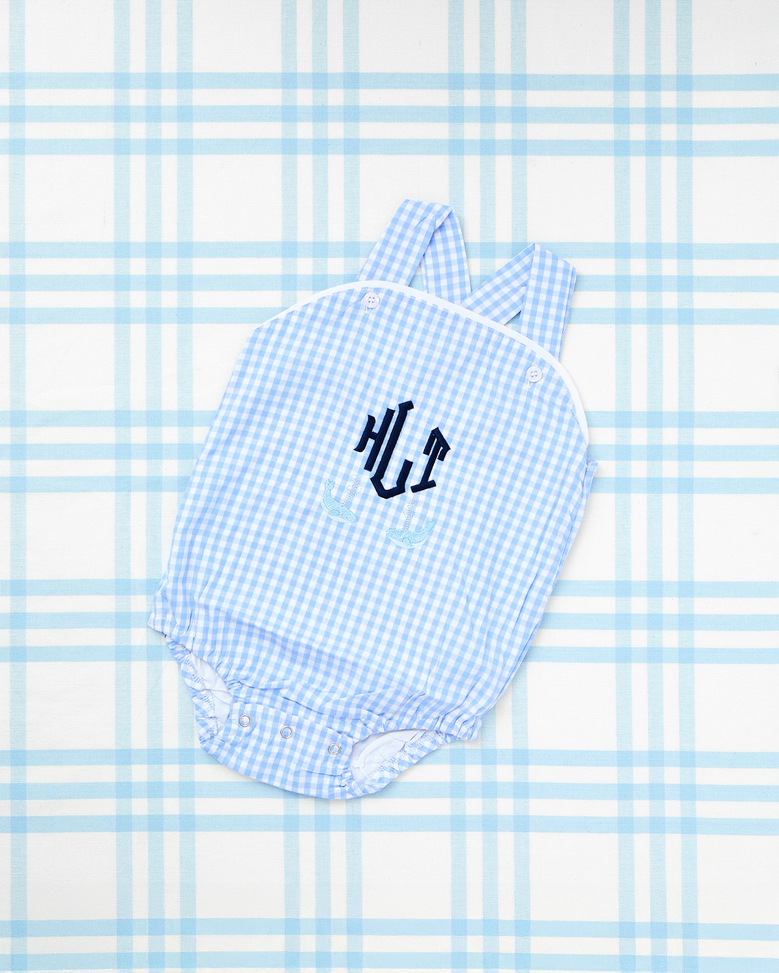 Image showcases two Gingham Sunsuits with Motif. The left sunsuit features a red gingham pattern with personalized navy blue initials and embroidered blue sailboats beneath, offering a nautical-inspired look. The right sunsuit displays a light blue gingham pattern with the name “Barrett” embroidered in red, complemented by a trio of colorful embroidered cars above the name. Both sunsuits include button closures and criss-cross straps, perfect for stylish and playful summer outfits.