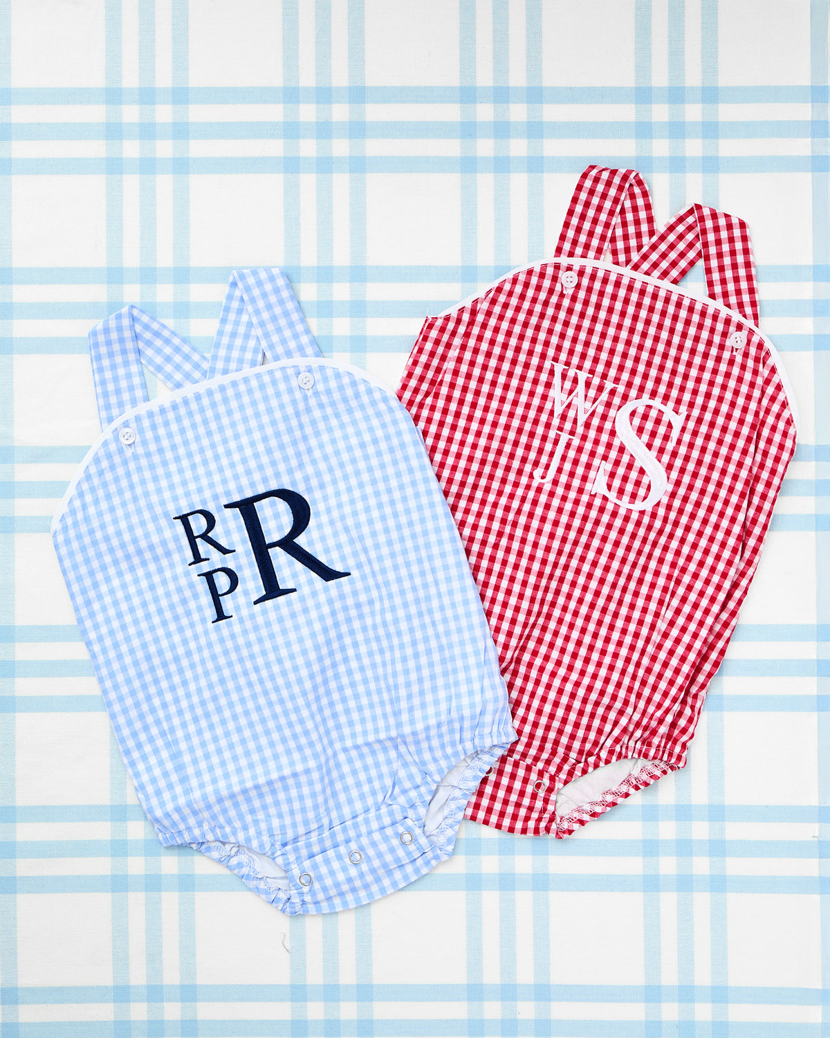 Flat lay of two classic Gingham Sunsuits for babies, displayed on a blue plaid backdrop. One sunsuit features a light blue gingham print with navy blue stacked monogram "RPR," while the other showcases a bold red gingham print with white monogram "YS." Both sunsuits include button detailing and criss-cross straps, offering a timeless and personalized look perfect for summer outings or Fourth of July celebrations.