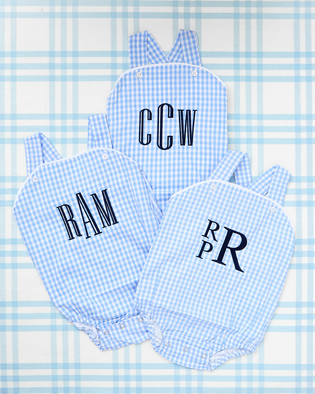 Flat lay of three personalized Gingham Sunsuits in light blue, arranged on a soft blue plaid backdrop. Each sunsuit features a unique navy embroidered monogram—"CCW," "RAM," and "RPR"—in classic fonts, adding a custom, timeless charm. These sleeveless sunsuits include white button accents and criss-cross straps, perfect for summer days or special occasions with a Southern-inspired, preppy flair.