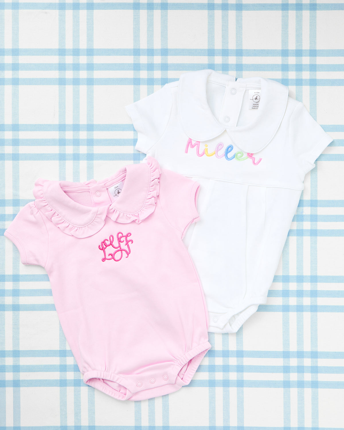 Flat lay of two Custom Name Short Sleeve Peter Pan Collar Bubbles displayed on a soft blue plaid background. The white bubble features the name “Miller” in a cheerful rainbow gradient embroidery, while the pink bubble showcases a ruffled collar and a bold hot pink monogram design. Both pieces highlight classic Southern charm and personalized style—perfect for everyday elegance or special milestone moments.