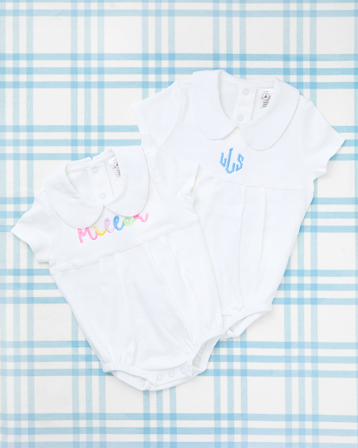 Flat lay of two elegant Custom Name Short Sleeve Peter Pan Collar Bubbles displayed on a soft blue plaid background. Each bubble features a classic white knit design with delicate pleats and signature rounded collars. One bubble is personalized with the name “Miller” in a playful multicolor embroidery, while the other showcases the initials “WJS” in a timeless baby blue thread. These personalized outfits combine comfort and charm, making them perfect for everyday wear or special occasions.