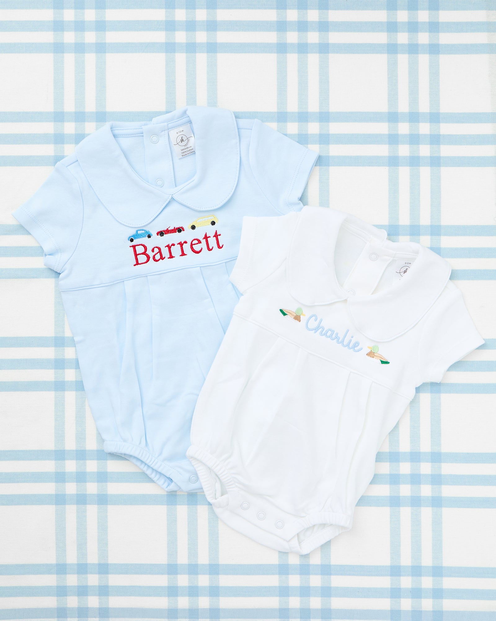 Image featuring two personalized Short Sleeve Peter Pan Collar Bubbles with Motif. The left outfit is a light blue bubble with a classic Peter Pan collar, showcasing embroidered golden retrievers flanking a row of red hearts and the name "MASON" in blue. The right outfit is a pastel pink bubble with a ruffled Peter Pan collar, adorned with dainty embroidered tulips and the name "Evelyn" in soft pink script. These adorable, monogrammed bubbles combine comfort and charm for any little one’s wardrobe.