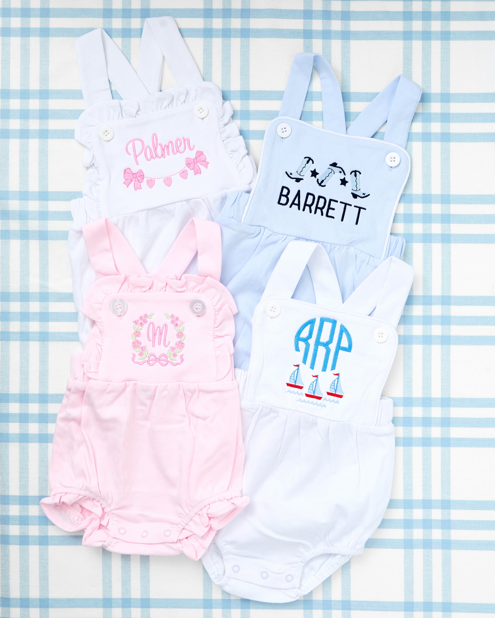 Image of two Cotton Sunsuits with Motif for toddlers—one in light blue and one in soft pink. The blue sunsuit features embroidered train cars above the name "KEEGAN," while the pink version showcases adorable puppy motifs and a string of hearts above the name "Sophia." Both sunsuits have shoulder button straps and elastic leg openings for a comfy, playful fit.