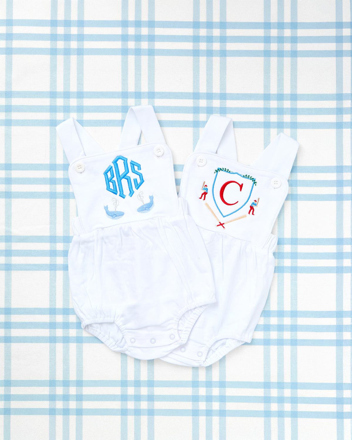 Two white cotton sunsuits with adjustable straps, each featuring custom embroidered motifs—one with whales and a monogram, and the other with a playful crossed bat and shield design—perfect for timeless, personalized baby style.