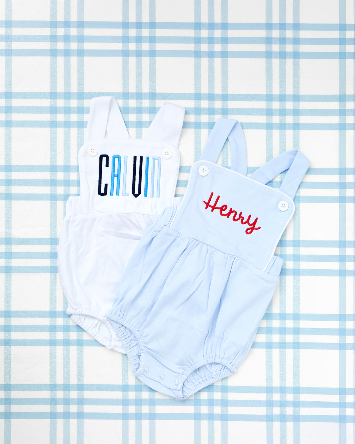 Image of two soft and stylish Cotton Sunsuits for babies, displayed on a blue gingham background. One sunsuit is white with “CALVIN” embroidered in bold blue gradient block letters, while the other is light blue with “Henry” stitched in bright red script. Both feature classic buttoned straps and elastic leg openings, blending comfort and personalized charm for everyday wear or special summer outings.