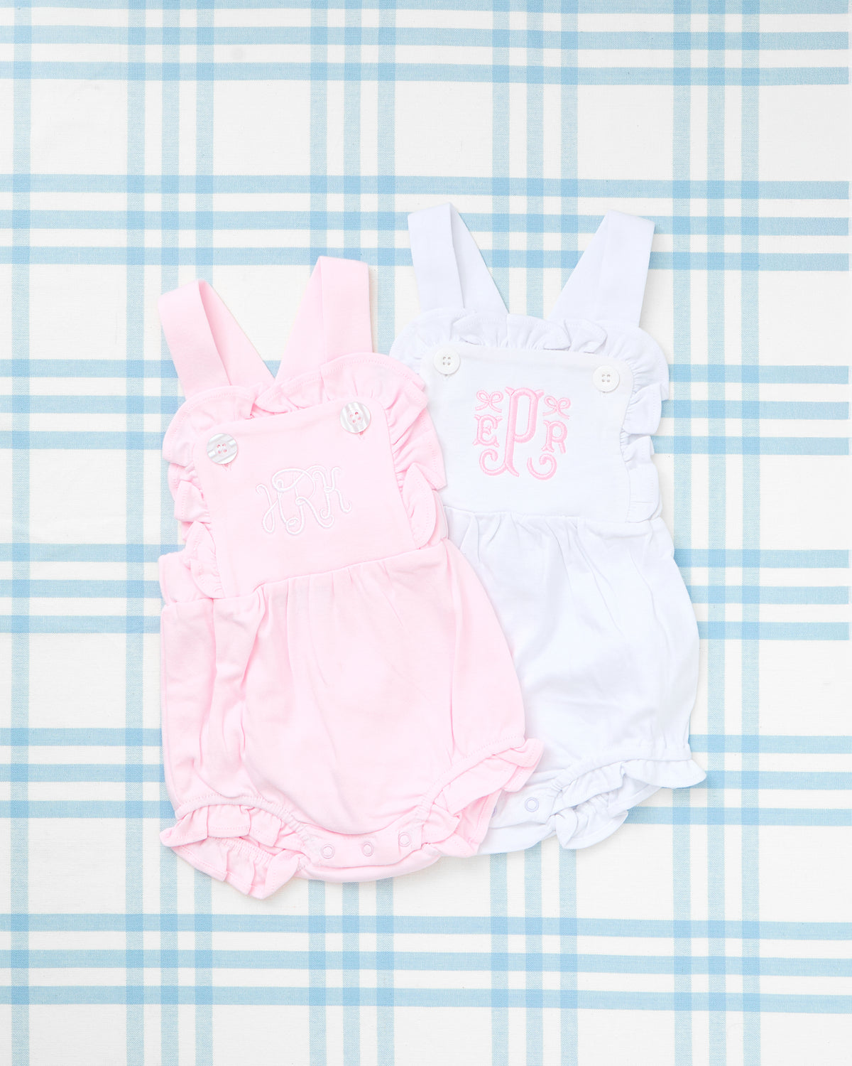 Image of two adorable Cotton Sunsuits laid out on a light blue gingham background. The pink and white sunsuits feature delicate ruffle trim, buttoned straps, and personalized embroidered monograms on the front chest panel, offering a charming and customizable outfit option for little ones. Perfect for warm-weather days and special occasions.