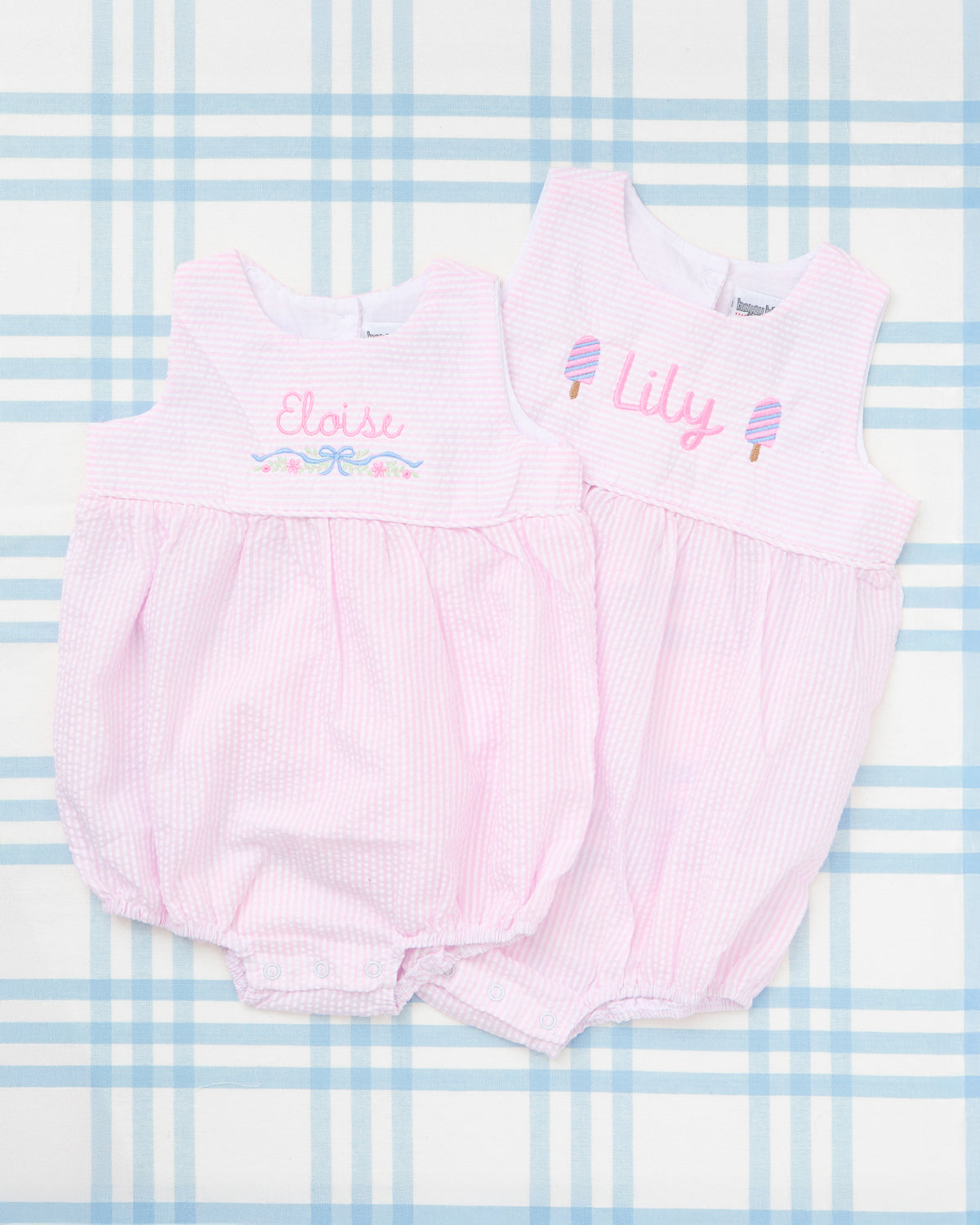 Flat lay of two pink Seersucker Bubbles with Motif displayed on a light blue plaid background. One bubble features the name “Eloise” with an embroidered floral bow motif, while the other showcases “Lily” with two embroidered ice cream cones. Both garments highlight sweet, summery personalization perfect for baby girls.