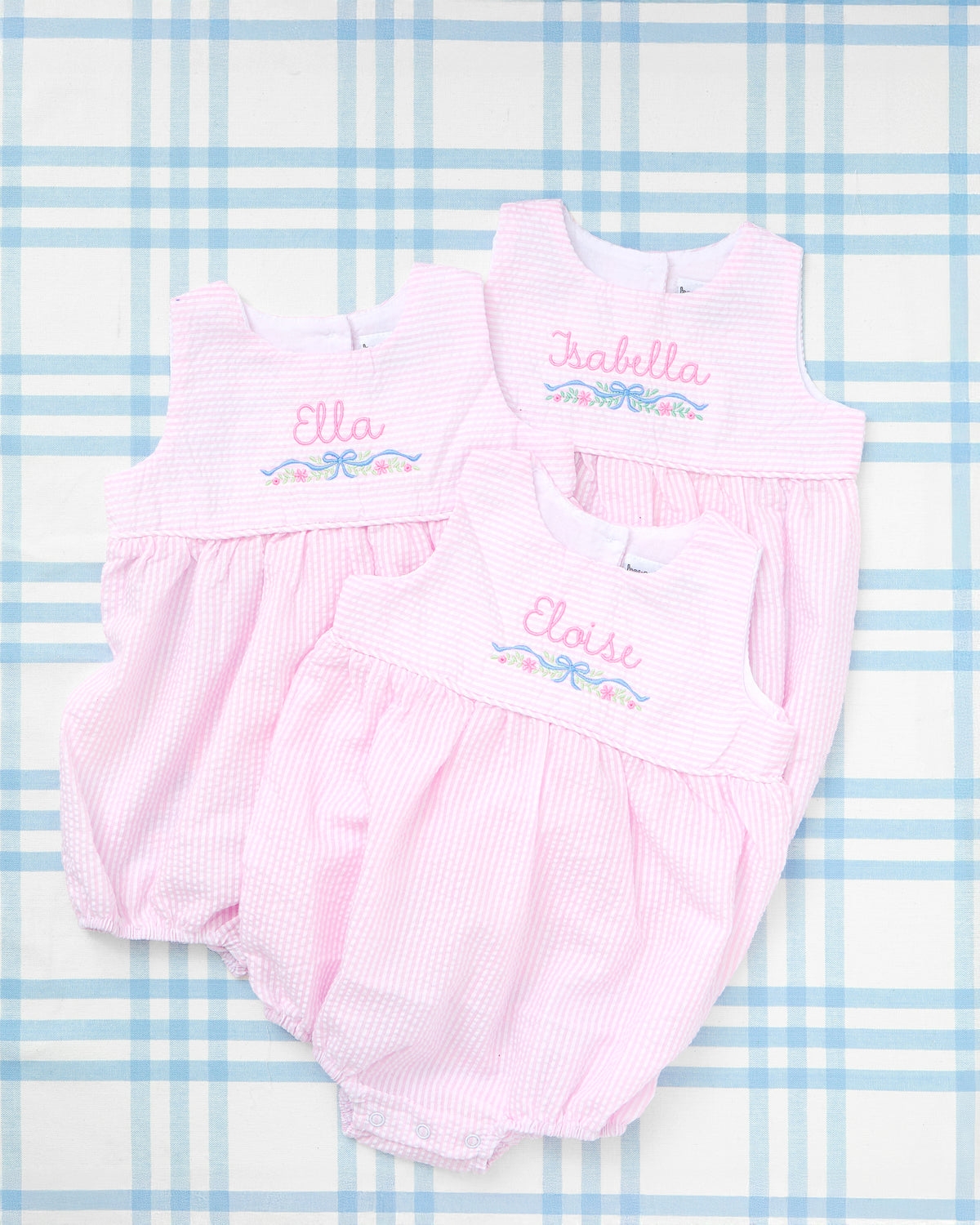 Three personalized Seersucker Bubbles with Motif in light pink, featuring delicate floral embroidery and names—Ella, Isabella, and Eloise—stitched in playful script across the chest. These charming one-piece outfits combine classic Southern style with sweet, custom details perfect for baby girls.