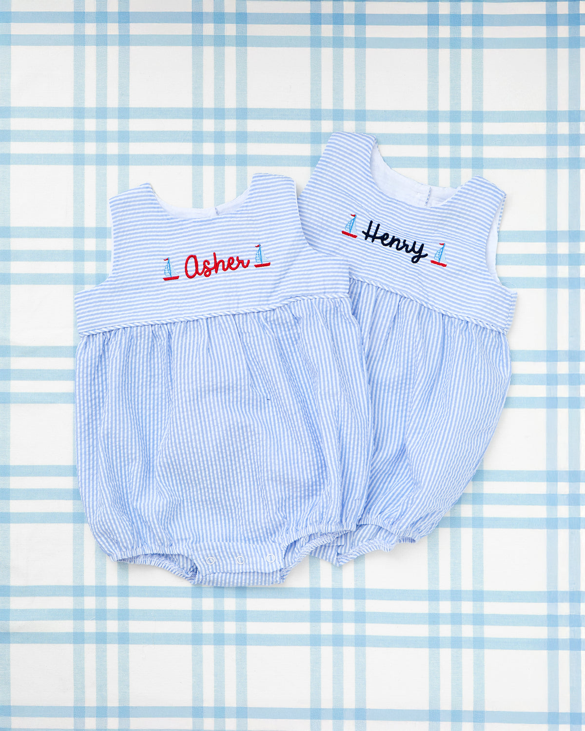 Flat lay of two personalized Seersucker Bubbles with Motif from Rainey Laine, made with soft blue and white striped fabric. Each bubble features a sailboat motif flanking embroidered names—“Asher” in red and “Henry” in navy. These classic, sleeveless baby outfits are ideal for summer celebrations and nautical-themed occasions.
