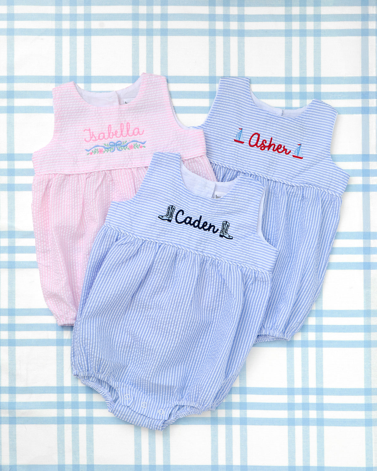 Flat lay featuring three Seersucker Bubbles with Motif from Rainey Laine, each personalized with a child’s name and unique embroidery design. The pink bubble for “Isabella” showcases a delicate floral motif with a pastel banner, while the blue bubble for “Asher” displays sailboat icons on either side of the name. The third bubble, in blue seersucker, features the name “Caden” embroidered between two cowboy boots. These classic, Southern-style outfits are perfect for summer photos and special occasions.