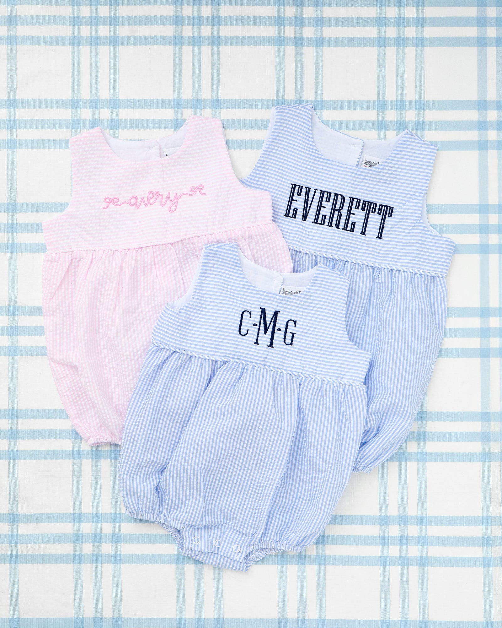 Personalized Seersucker Bubbles from Rainey Laine featuring classic sleeveless designs—one in light blue with navy monogram ‘JBH’ and one in pink with bright pink monogram ‘EMG’—crafted in soft striped fabric with gathered bottoms for a timeless baby look.