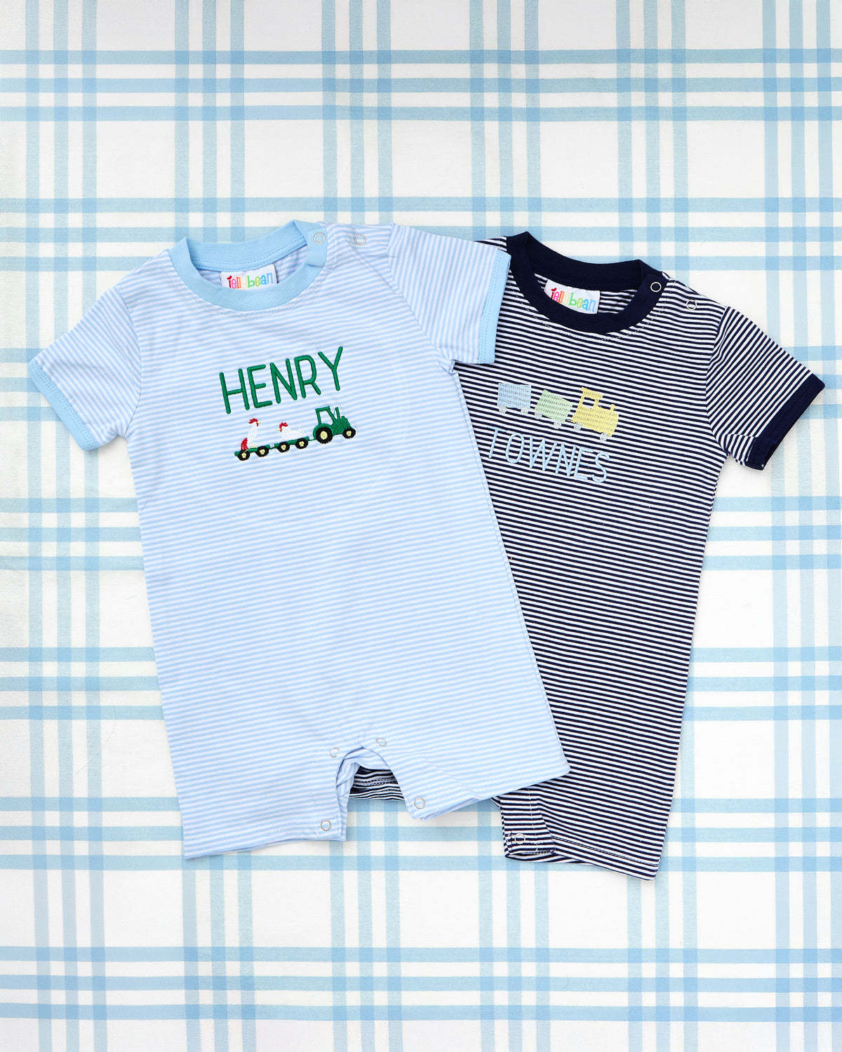 Striped Short Sleeve Romper & T-Shirt with Full Chest Motif from Rainey Laine, featuring a light blue romper with a green tractor and trailer motif personalized with the name ‘Henry,’ and a navy romper with a yellow bulldozer and dump truck motif customized with the name ‘Townes,’ both displayed on a blue plaid background.