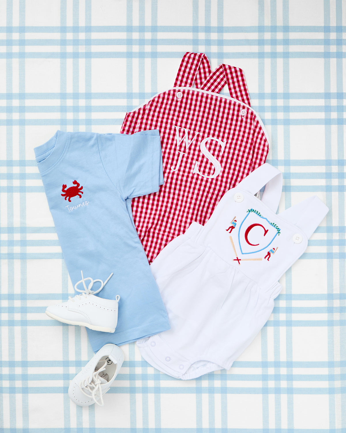 Flat lay of a vibrant red Gingham Sunsuit with white monogram initials “Y S,” displayed alongside a light blue embroidered t-shirt featuring a red crab and name detail, and a white cotton sunsuit with a red-and-blue Baseball Crest motif and initial “C.” Set on a soft blue plaid background with white baby shoes, this summery outfit trio exudes preppy charm and coastal playfulness—perfect for personalized warm-weather looks.