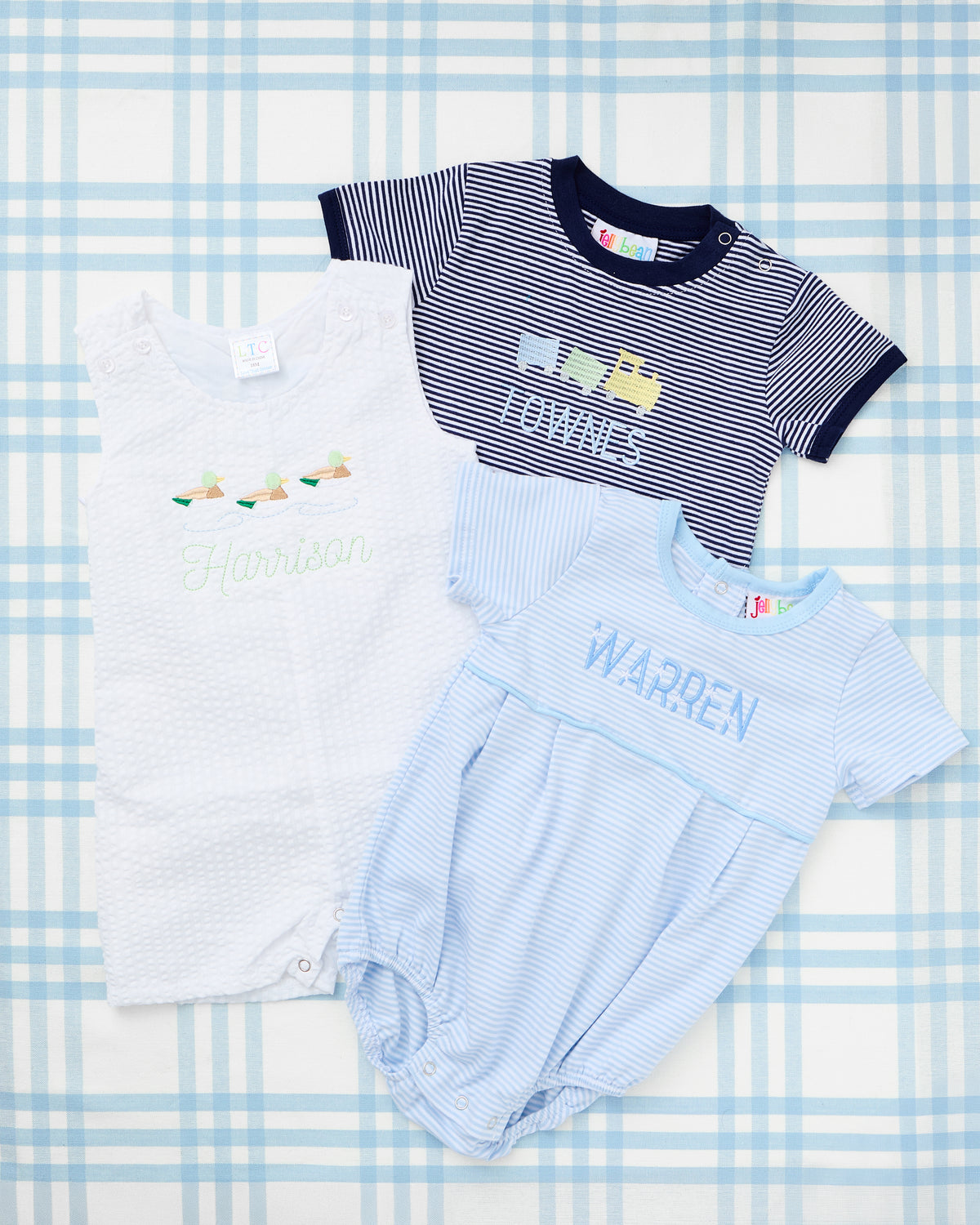 Short Sleeve Striped Bubbles from Rainey Laine featuring personalized designs—light blue striped bubble with a green tractor trailer motif and name ‘Henry,’ and navy striped bubble with yellow construction truck motif and name ‘Townes,’ both laid out on a soft blue plaid background.